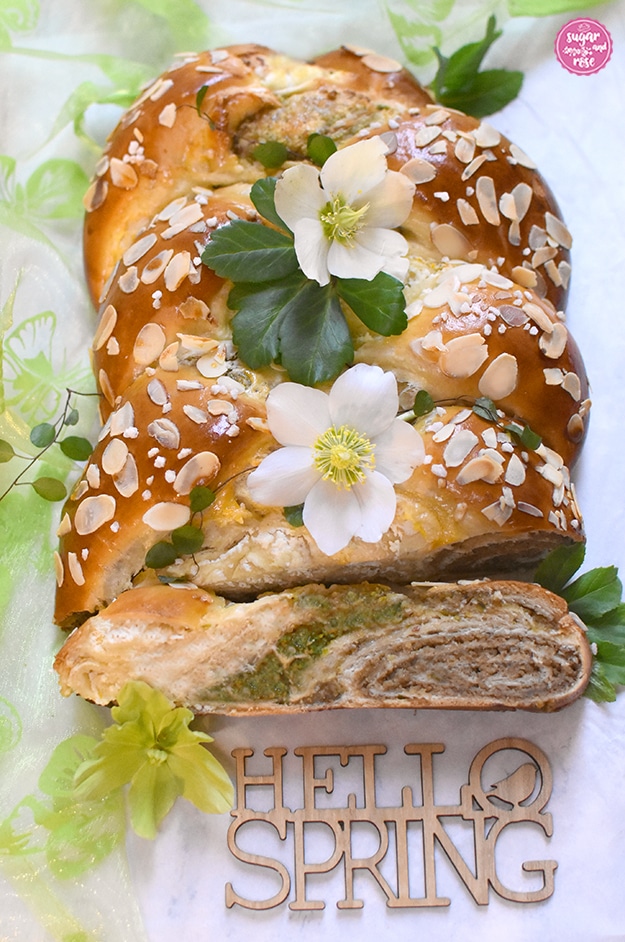 Angeschnittener Briochestriezel (Hefezopf), dekoriert mit weißen Schneerosenblüten und grünen Blättern, im Vordergrund ein Stück Striezel, wo man die drei Füllungen sieht: Topfen links, mittig die Pistazien, rechts die Nüsse. Davor ein Schriftzug aus Sperrholz "Hello Spring".