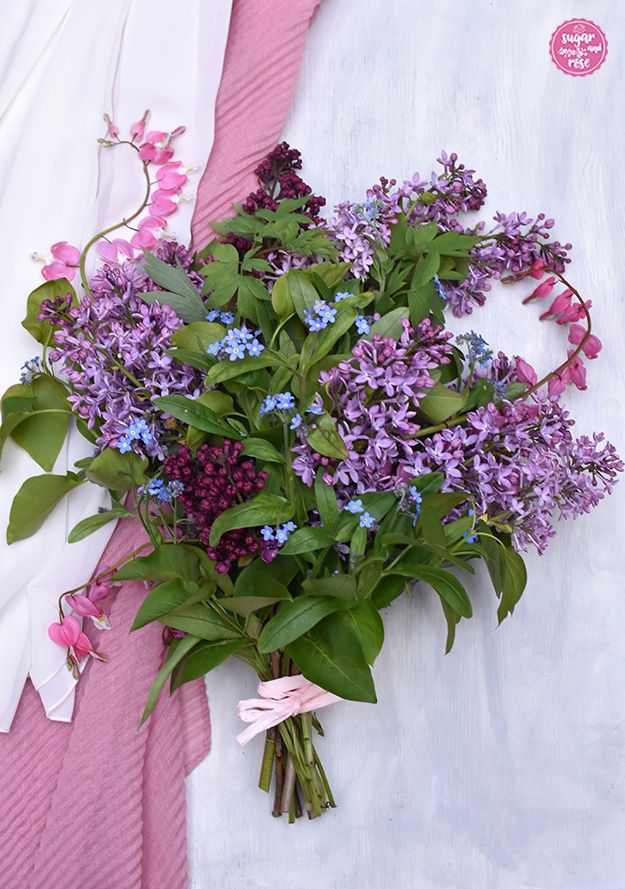 Muttertagsblumenstrauß aus Flieder, Vergissmeinnicht und Herzblumen mit einem hellrosa Bändchen zusammengebunden, auf einem weißen Untergrund mit rosa Tuch liegend.