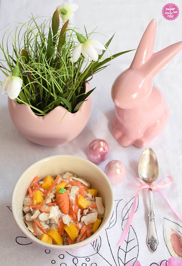 Auf einem Tischtuch mit österlichem Hasenmotiv steht vorne ein rosa Keramikschälchen mit Overnight Oats und Karotten und Mango, daneben ein Siberlöffelchen mit rosa schleife, dahinter ein rosa Porzellanhase, zwei rosa glitzernde Porzellaneier und eine rosa Vase in Form eines halben Eis, darin grünes Gras und drei Frühlingsknotenblumen