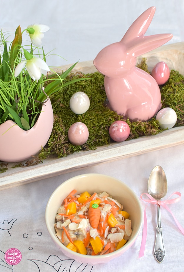 Rosa Keramikschälchen mit Overnight Oats und Karotten und Mango, daneben ein Siberlöffelchen mit rosa schleife, dahinter eine weiße Holzschale mit rosa Porzellanhase, perlmuttschimmernde rosa Porzellaneier und eine rosa Blumenschale in Form eines zerbrochenen rosa Eis