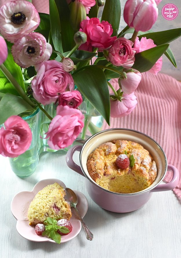 Hirse-Topfenauflauf mit Himbeeer-Garnitur im rosa Reindl, davor ein blütenförmiger rosa Teller mit einem Stück Auflauf, Himbeeren und einem silbernen Teelöffel, dahinter die Aalto-Vase mit rosa Tulpen und Ranunnkeln.