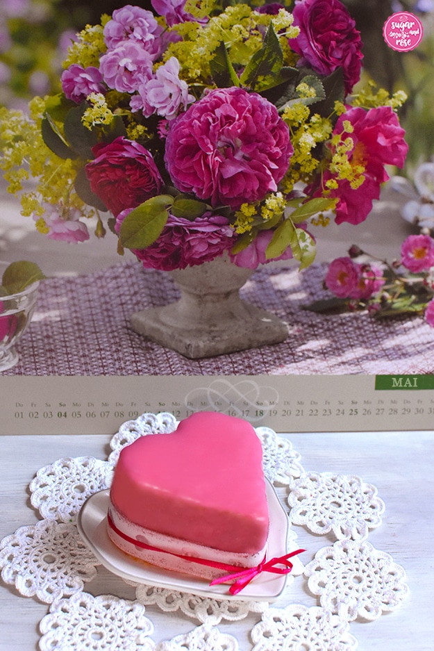 Kalenderblatt "Mai" mit rosa Rosen in einer Steinvase, davor auf einem gehäckelten weißen Deckchen ein Punschkrapfenherz auf einem herzförmigen Teller