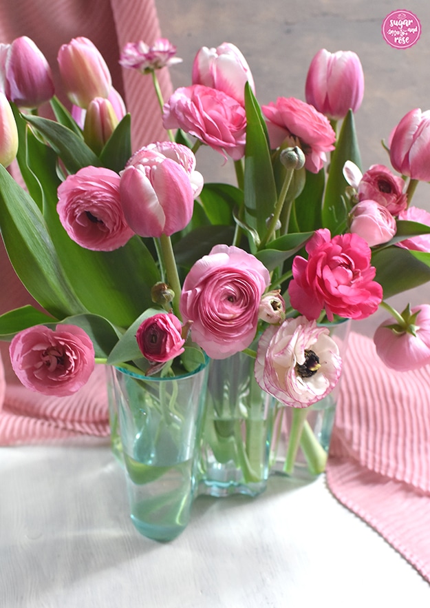 Eine mittelgroße durchsichtige grüne Aalto-Vase im klassischen skandinavischen Design mit einem dichten Strauß an rosa-weißen Tulpen und Ranunkeln in verschiedenen Rosatönen, im Hintergrund ein pastellrosa Tuch mit Faltenwurf.