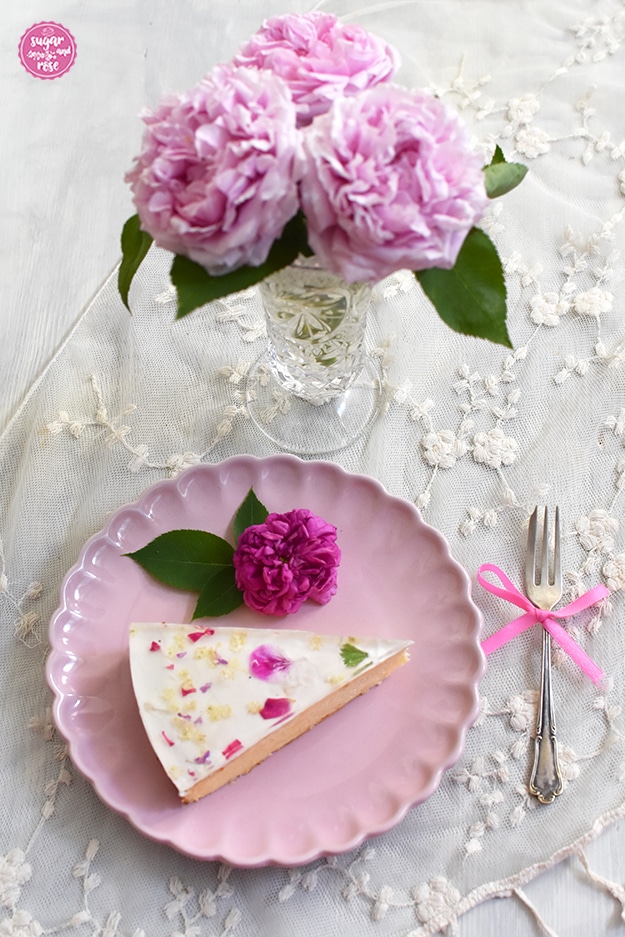 Ein Stück Topfentorte mit Blütem auf einem rosa Porezellanteller, daneben eine Vintagekuchengabel mit rosa Schleife. Im Hintergrund eine Glasvase mir rosa Rosen.