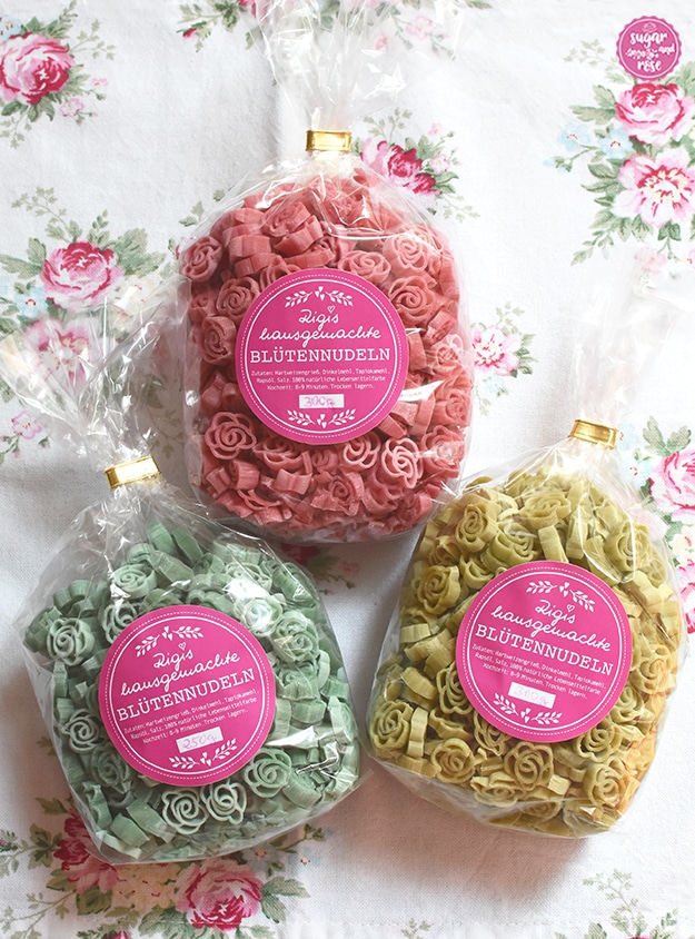 Drei Packungen Blütennudeln: rosafarbene, hellgrüne und mintgrüne Rosennudeln in Cellophanpackungen mit rosa Aufkleber
