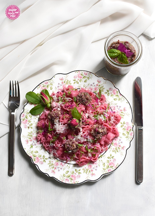 Rosendekorteller mit Rigis Rosennudeln mit Rosenpesto, daneben Edelstahlbesteck und ein weißes Tuch sowie ein Glas mit Rosenpesto