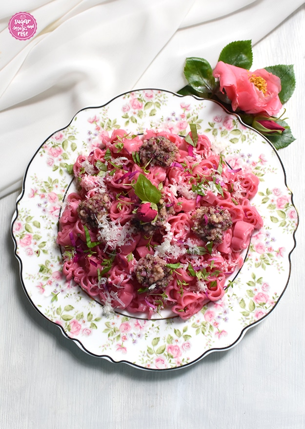 Rosafarbene Blütennudeln mit Rosenpesto auf einem Teller mit Rosendekor, daneben eine lachsfarben Kamelienblüte