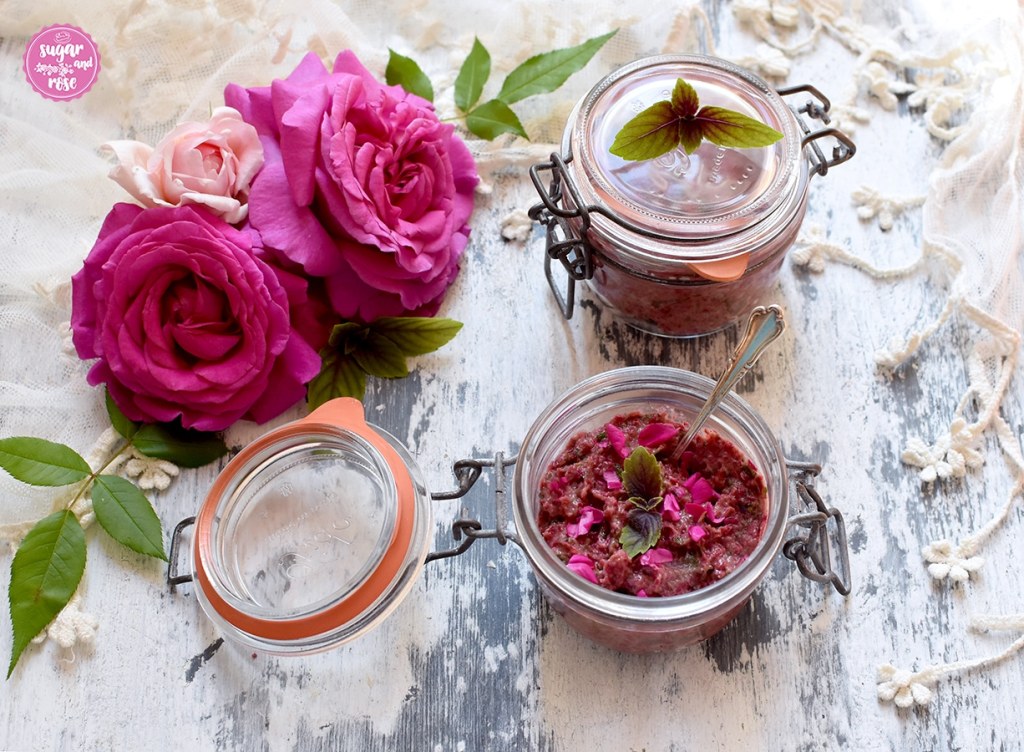 Rosenpesto in Rexgläser mit Deckel gefüllt, ein Glas offen mit einem Teelöffel eingesteckt, im Hintergrund ein cremeweißes Spitzentuch und zwei pinkfarbene und eine blassrosafarbene Rose