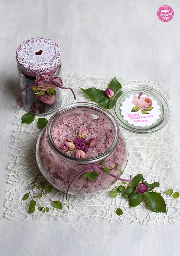 Ein großes bauchiges Rexglas gefüllt mit Rosen-Body-Peeling, dekoriert mit kleinen getrockneten rosa Rosenknospen, dahinter ein Glas mit getrockneten Rosenblüten und der Deckel des Glases mit Beschriftung in Rosa