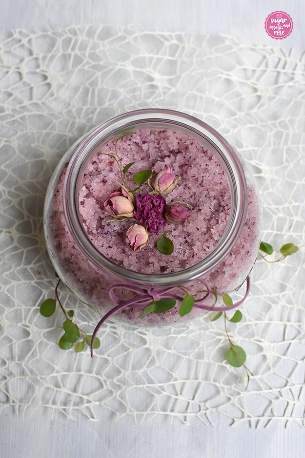 Offenes Rexglas von oben, man sieht das körnige rosa Peelingsalz, das Glas steht auf einem weißen netzförmigen Deckerl und ist mit grünen kleinen Blättern und eine rosa Masche dekoriert