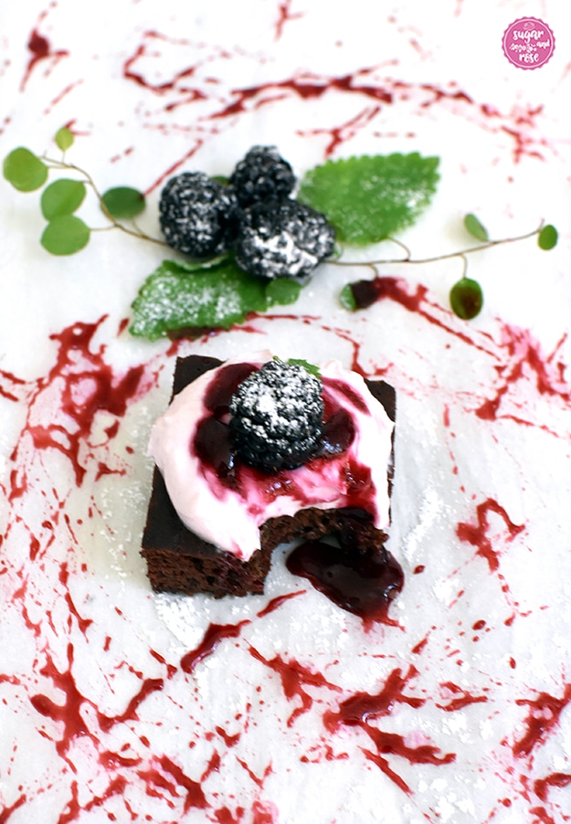 Kalorienarmer Brownie mit Frischkäsegarnitur und viel dunkelroter Brombeersauce verteilt am weißen Untergrund, dahinter drei dunkelschwarze große Brombeeren mit Puderzucker und Minzblättern