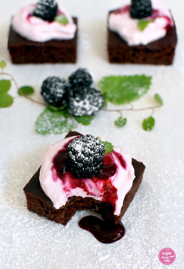 Drei kalorienarme Brownies mit Frischkäsetopping und Brombeeren, der Brownie im Vordergrund ist angebissen und es rinnt eine dicke Schicht bordauxfarbene Brombeersauce über den Brownie