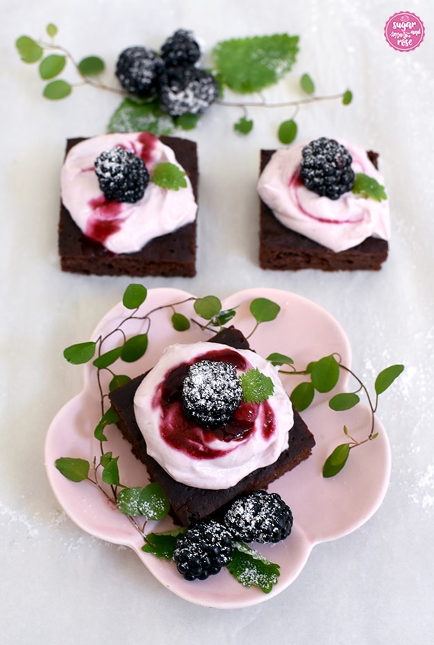 Ein Brownie auf einem rosa blütenförmigen Keramiktellerchen, dekoriert mit Brombeeren und grüne Blättern, dahinter zwei Brownies mit lilafarbenem Frischkäsetopping und Brombeeren