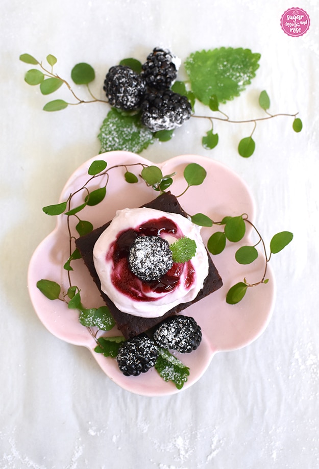 Ein Brownie auf einem rosa blütenförmigen Keramiktellerchen, dekoriert mit Brombeeren und grüne Blättern
