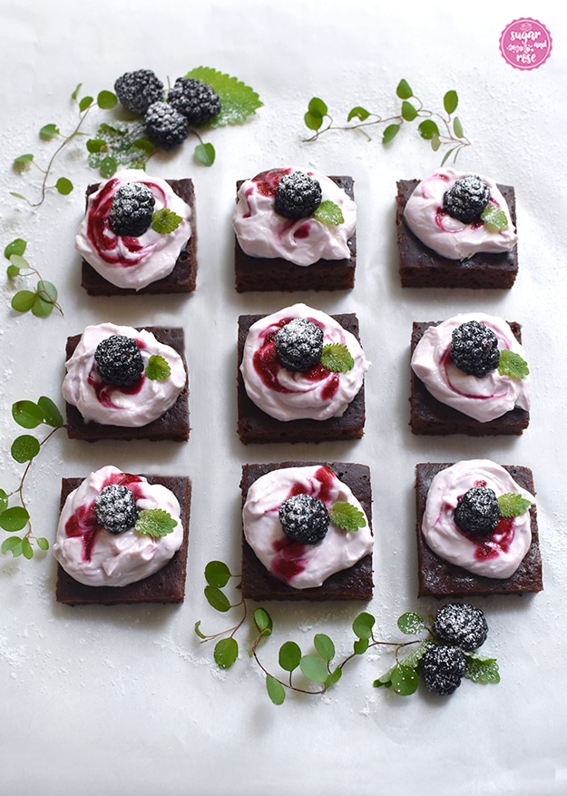 9 Stück kalorienarme Brownies mit Frischkäsetopping in Dreierreihen aufgelegt, dahinter frische Brobeeren und grüne Blätter