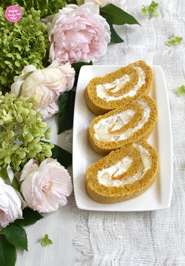 Drei Stück Kürbisroulade gefüllt mit Birnen und Mandelcreme auf weißem rechteckigen Porzellanteller, daneben grüne Hortensienblüten und blassrosa Rosen
