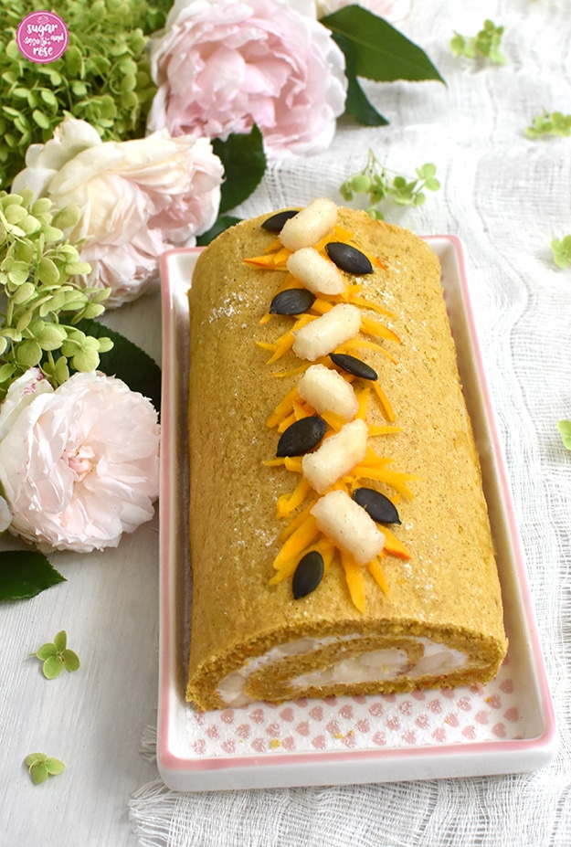 Kürbisroulade auf rechteckigem Porzellanteller mit rosa Herzchen-Muster, dahinter drei blassrosa Rosen und grün-weiße Schneeballhortensie