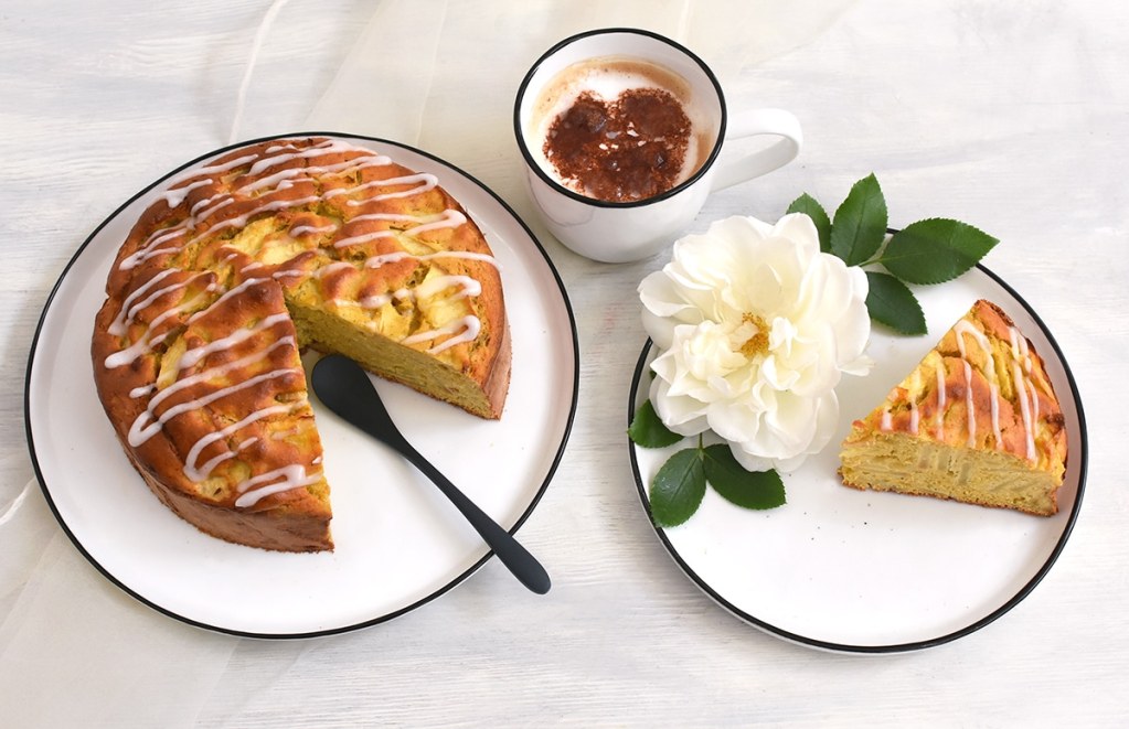 Leichter Apfelkuchen mit Kichererbsenmehl auf weißem rundem Teller mit dunkelblauem Rand, davon ein großes Stück herausgeschnitten auf eigenem Teller, dekoriert mit großer weißer Rosenblüte, dahinter eine Schale Melange mit Kakaoherz