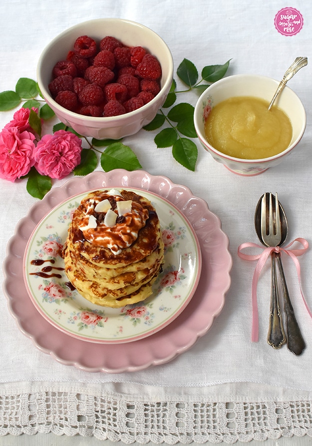 Pancakes auf Blütenteller und rosa Unterteller, daneben Silbergabel und Löffel mit rosa Schleife, dahinter eine Schale mit Himbeeren und eine mit Apfelmus, dekoriert mit rosa Rosen
