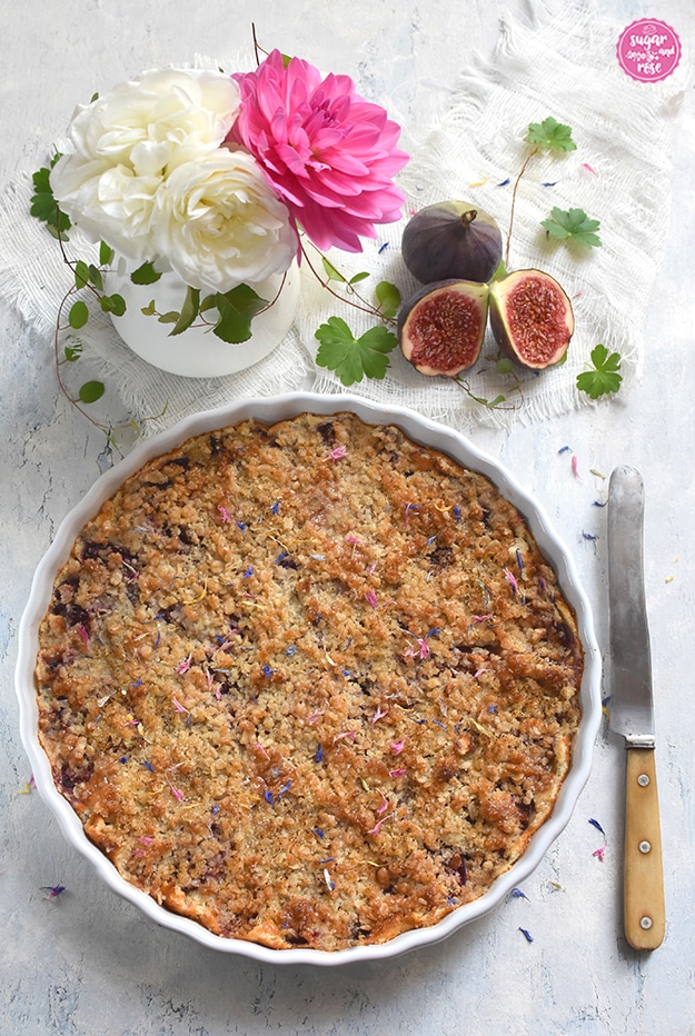 Zwetschken-Feigen-Streuselkuchen in weißer runder Keramiktarteform, daneben aufgeschnittene Feigen und ein altes Messer mit hellbraunem Griff, daneben ein weißer Krug mit weißen Rosen und einer pinken Dahlie