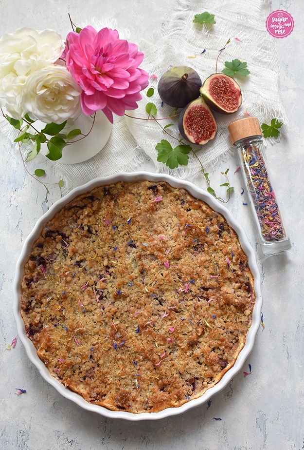 Zwetschken-Feigen-Streuselkuchen in weißer runder Keramiktarteform, daneben aufgeschnittene Feigen, eine Glasphiole mit getrockneten bunten Blüten und ein weißer Krug mit weißen Rosen und einer pinken Dahlie