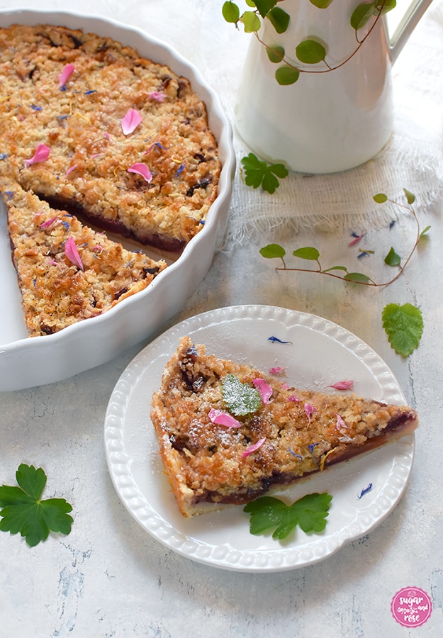 Ein Stück vom Zwetschen-Feigen-Streuselkuchen auf einem kleinen weißen Porzellanteller im Vordergrund, hinten die weiße Keramiktarteform mit dem Kuchen