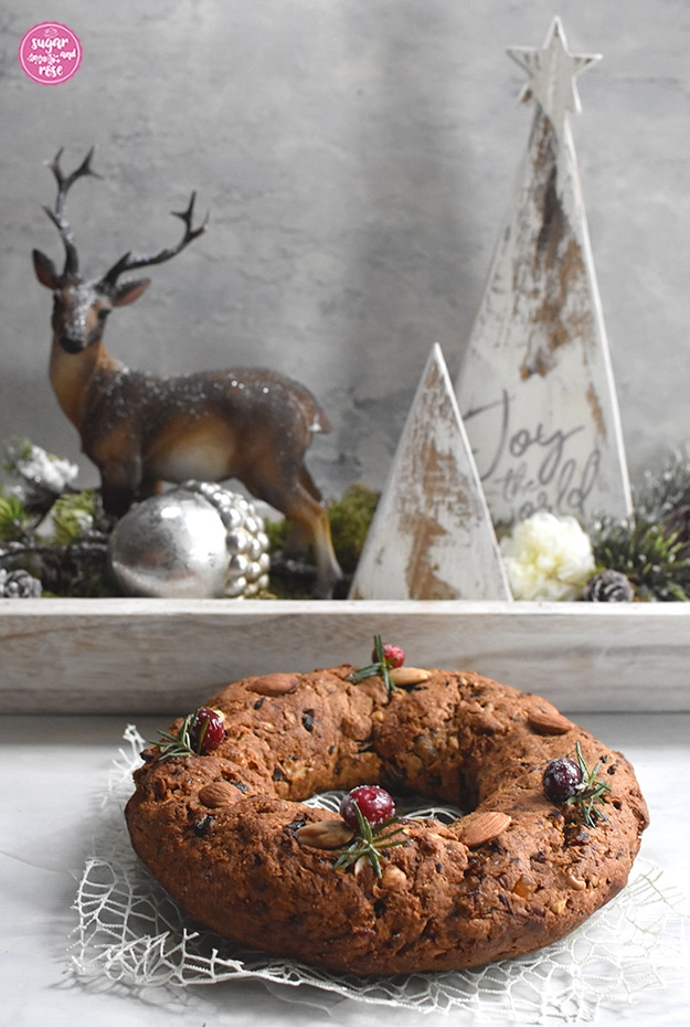 Kletzenbrot in Ringform, dekoriert mit Cranberrys, Rosmarin und Mandeln vor einer weißen Holzschale mit Hirschfigur und weißen Holzbäumchen