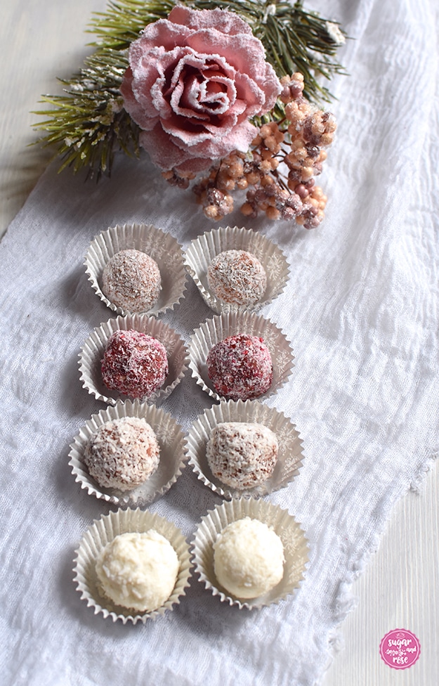 Weihnachtliche Energiekugeln in Papiermanschetten, je zwei Sorten nebeneinander in Viererreihe, dahinter eine rosa Kunstblumenrose und rosa Beeren mit Kunstschnee
