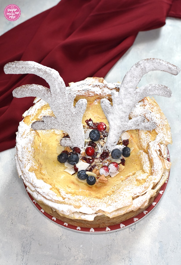 Spekulatius-Cheesecake mit weiß gezuckertem Rentiergeweih, Heidelbeeren und Preiselbeeren sowie Nüssen als Topping, dahinter ein weinroter Seidenschal