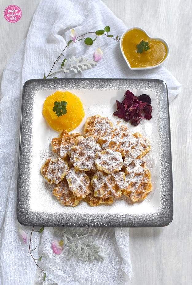 Topfenwaffeln auf einem rechteckigen weißen Keramikteller mit grauem Rand (Gmundner Keramik), daneben Orangenfilets und Rosenblüten, dahinter eine winzige herzförmige Porzellanschale mit Mango-Apfelmark