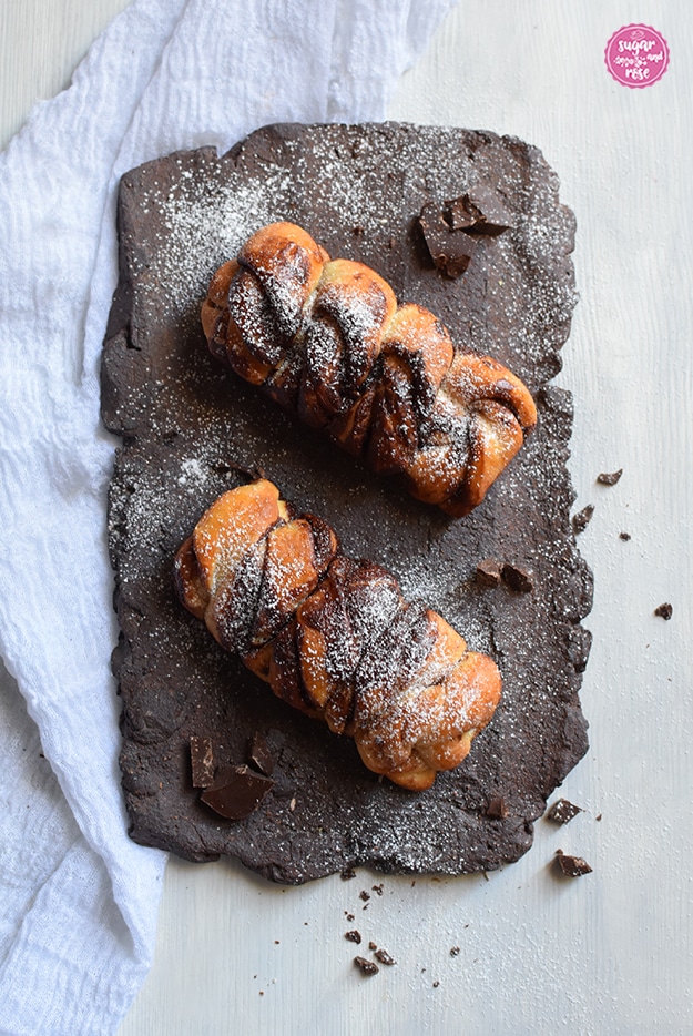 Zwei Schoko-Swirl-Heferollen auf dunkelbraunem dachziegelartigem rauem Teller, bestreut mit Puderzucker
