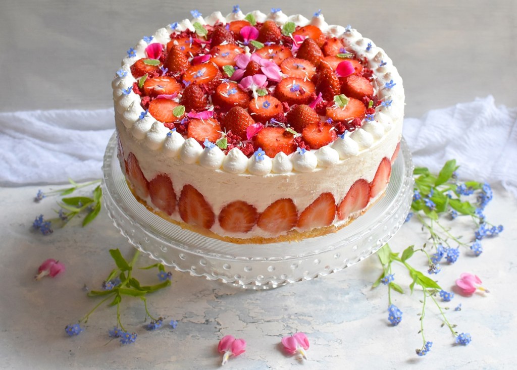 Erdbeer-Holundertorte mit weißer Creme und aufgeschnittenen Erdbeeren als Topping, dekoriert mit Vergissmeinnichtblüten und Minzblättern