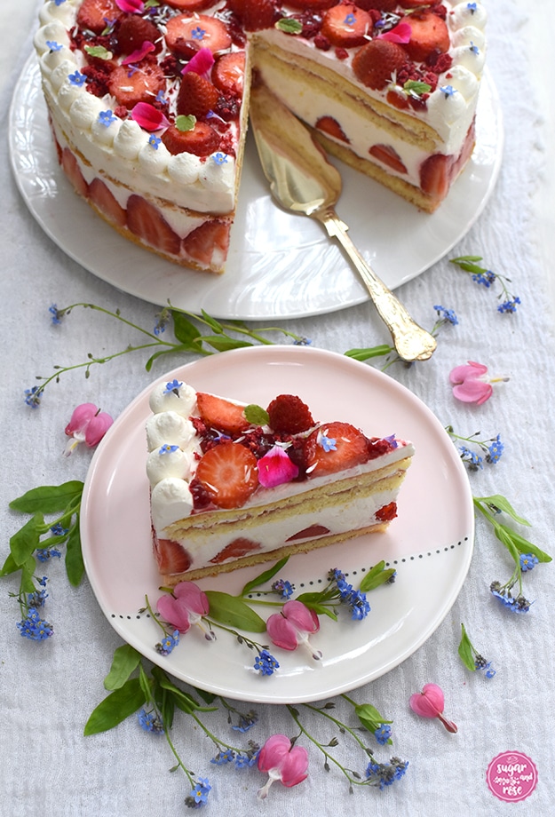 Im Hintergrund die angeschnittene Erdbeer-Holunder-Torte und ein kleiner silberner Tortenheber, davor ein rosa Dessertteller mit einem Stück der Torte, dekoriert mit Vergissmeinnicht und Blüten vom Herzerlstock