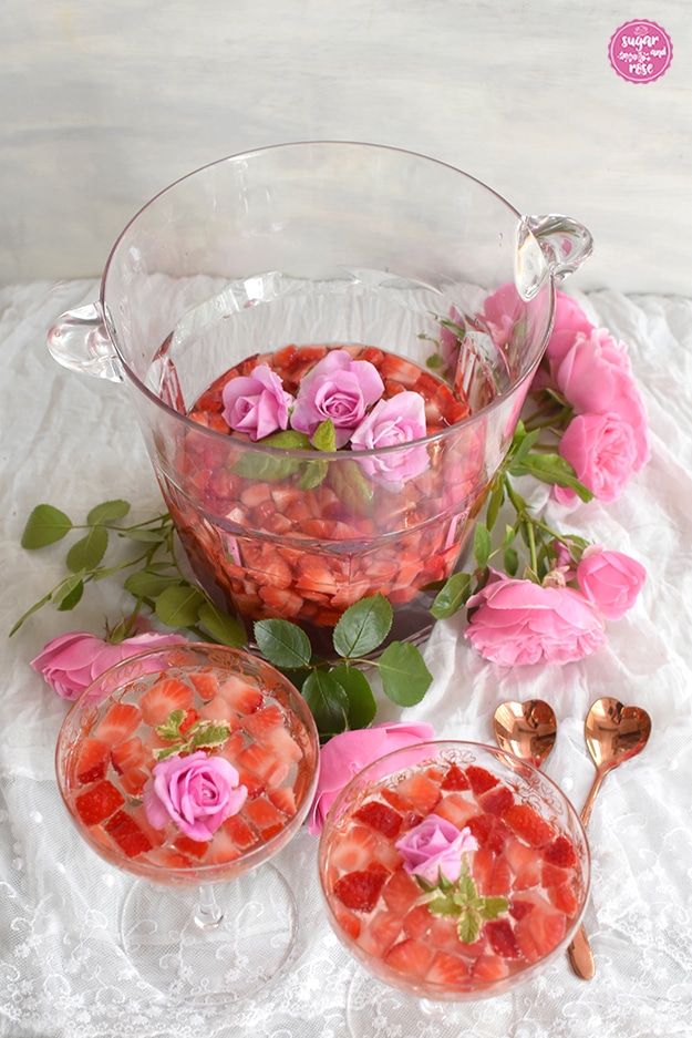 Durchsichtiger Sektkübel mit Erdbeeer-Rosenbowle, dekoriert mit drei sehr kleinen rosa Röschen, davor zwei Sektschalen mit Erdbeerbowle, zwei herzförmige bronzefarbene Löffelchen und rosa Rosen als Deko