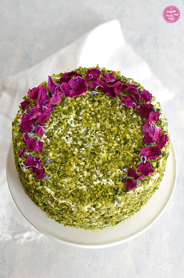 Pistazientorte von oben fotografiert, bedeckt rundum mit gehakten grünen Pistazien, am oberen Rand entlang hufeisenförmig mit getrockneten dunkelpinken Rosenblüten garniert