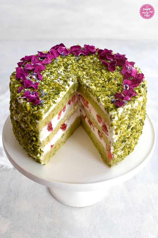 Pistazientörtchen auf weißer Etagere mit Fuß mit Anschnitt. Es fehlt ein Viertel der Torte. Sichtbar sind die drei weißen Cremeschichten mit Erdbeerstückchen