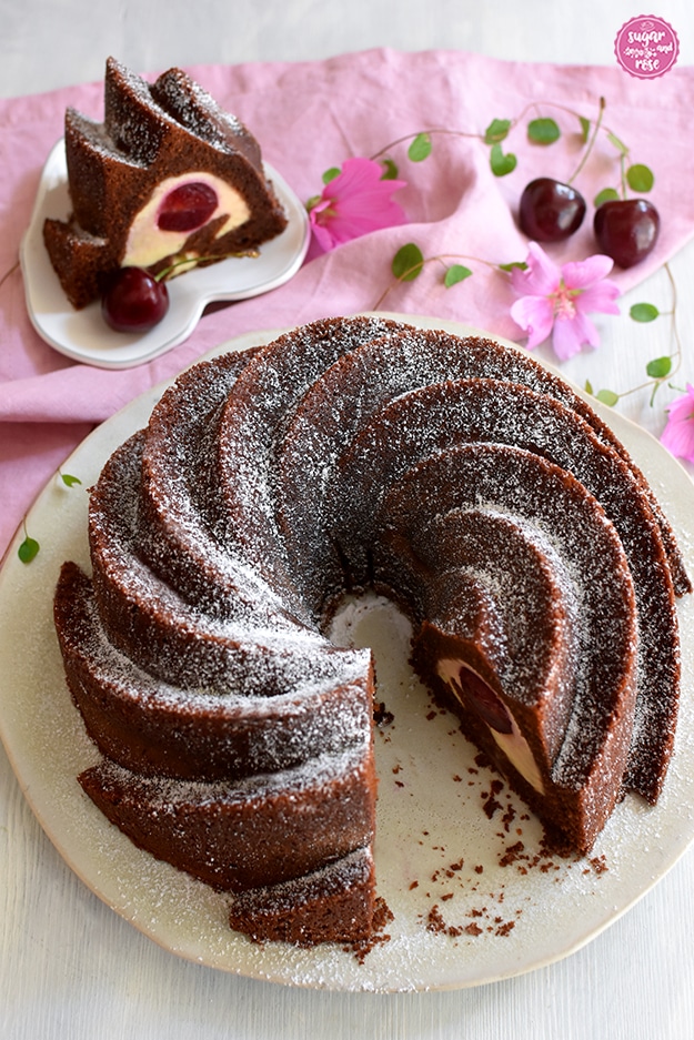 Angeschnittener Schoko-Kirschgugelhupf auf Keramikteller, dahinter ein herzförmiger Teller mit einem Stück Kuchen, man sieht die helle Topfenmasse innen und die Kirschen