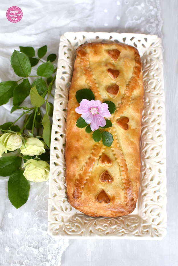 Ungezuckerte Apfelschlangerl, dekoriert mit Mürbteigherzchen auf durchbrochener Keramikschale, daneben grün-weiße Rosen.