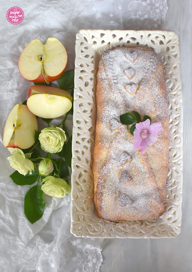 Apfelschlangerl auf Keramiktablett, dekoriert mit einer lila Malvenblüte, daneben ein aufgeschnittener Apfel und kleine grün-weiße Röschen.Bild rechts: ein Stück mürber Apfelstrudel und eine Tasse Kaffee, dahinter eine mintgrüne Keramikkaffeekanne