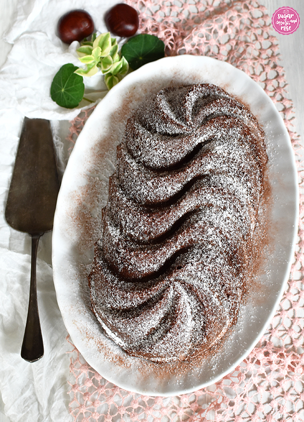 Maroni-Gewürzkuchen mit Staubzucker und etwas Kakao bestreut auf ovaler Platte, daneben ein alter Tortenheber, Kastanien und grüne Blätter