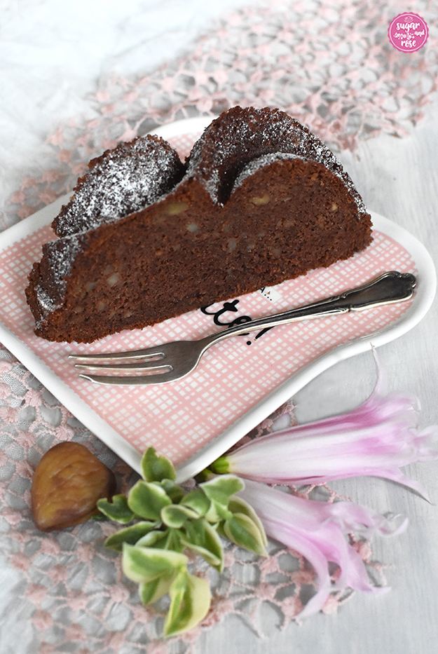 Je ein Stück Maroni-Gewürzkuchen auf einem kleinen toastbrotförmigen weiß-rosa-kariertem Kermamiktellerchen, daneben eine alte Kuchengabel, Maroni, grüne Blätter rosa Blüten