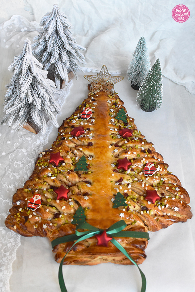 Hefe-Weihnachtsbaum mit Nussfüllung, dekoriert mit grüner Schleife und roten Sternen, im Hintergrund zwei kleine schneebedeckte Dekoweihnachtsbäume auf Holz und weiße Spitzendecke