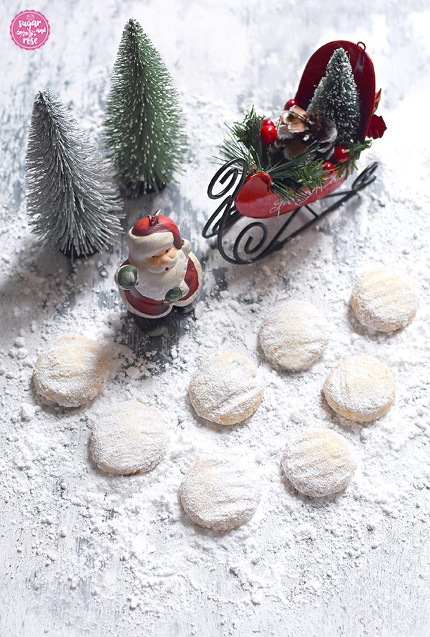 Einige gezuckerte runde Vanille-Cookies auf Holzbrett mit viel Staubzucker darunter; im Hintergrund ein kleiner Keramikweihnachtsmann, zwei Dekobäume und ein roter Weihnachtsmannschlitten