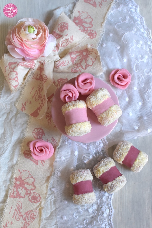 Hell- und dunkelrosa Punschrollen auf weißem Holzuntergrund und weißer Spitzendecke, zwei Rollen liegen mit zwei rosa Marzipanrosen auf einem winzigen rosa Teller, im Hintergrund eine rosa Ranunkelblüte und eine weiß-rosa Schleife