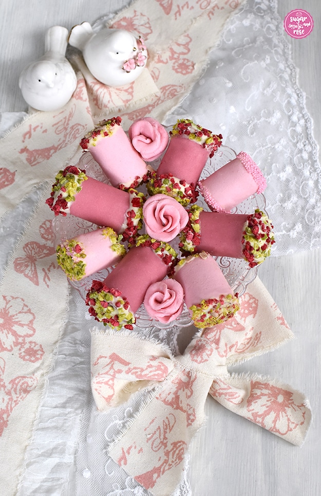 Hellrosa und dunkelrosa Punschrollen auf kleiner Glasetagere, dazwischen rosa Marzipanröschen und im Hintergrund zwei weiße Porzellantäubchen
