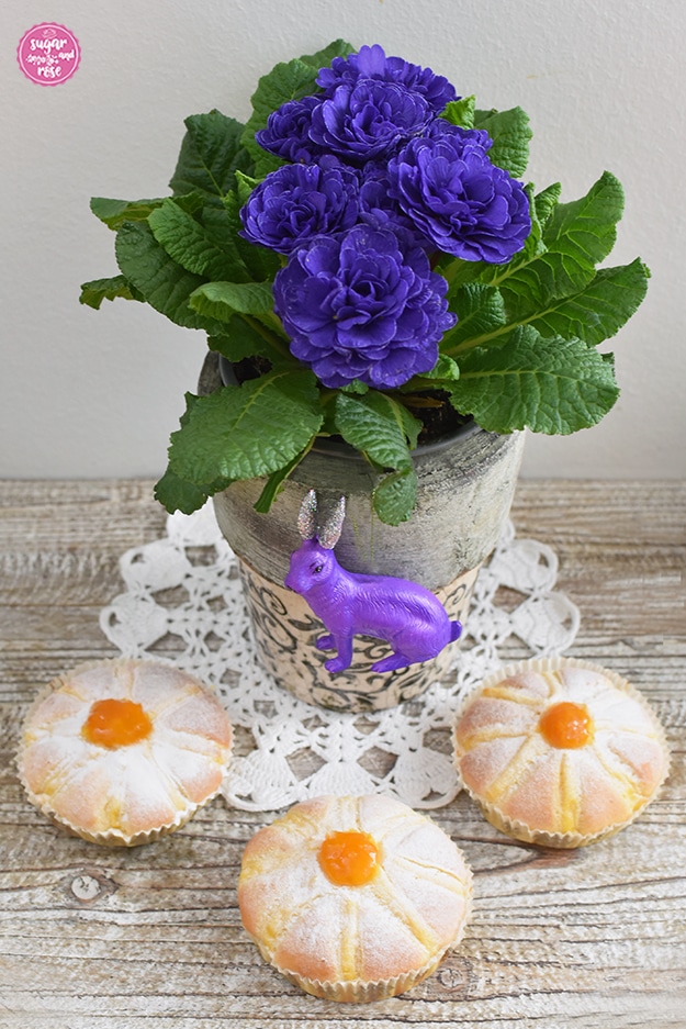 Drei Blüten-Tartelettes in Papiermanschetten vor eine gefüllten nahezu blauen Primel auf einem weißen Spitzendeckchen