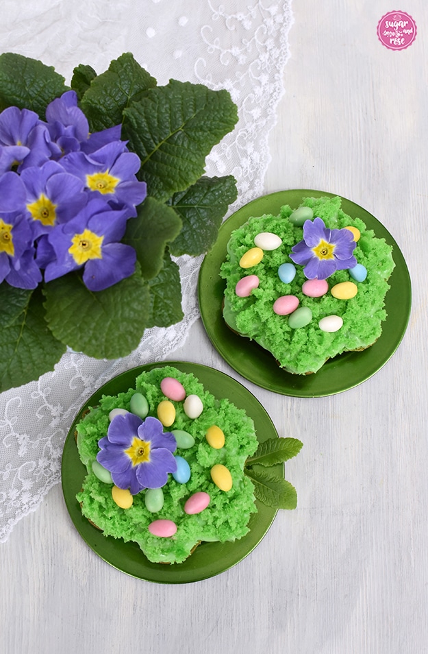 Zwei Mooskuchen-Osternester auf grünen Tellerchen, dekoriert mit je einer lila Blüte und pastellfarbenen Zuckerostereiern. Im Hintergrund eine lila Primel.