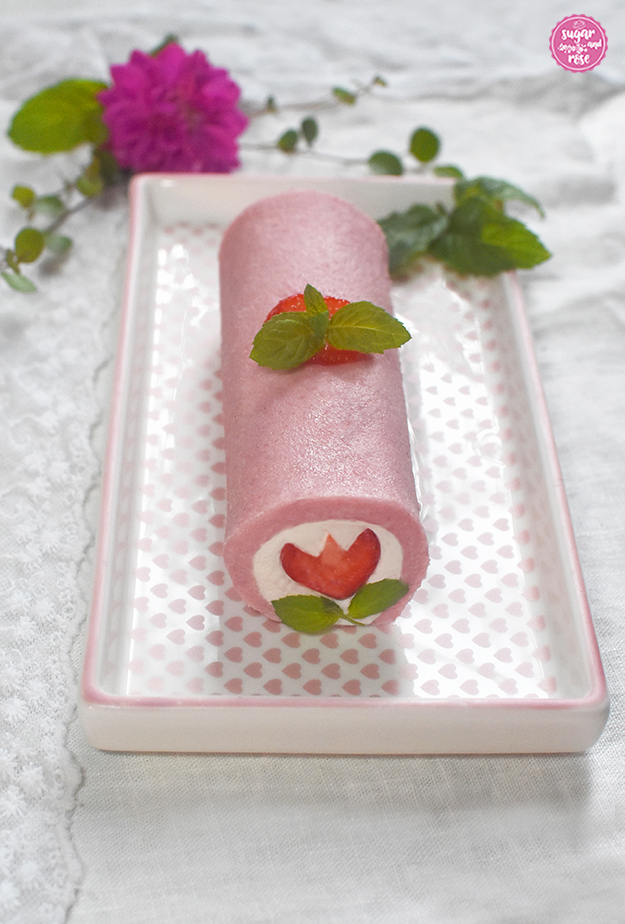 Rosa Biskuitrolle gefüllt mit weißem Obers, an der Schittkante in zu einer Blüte geschnittene Erdbeere mit zwei grünen Minzblättchen; die rosa Roulade liegt auf einem länglichen Teller mit zarten rosa Herzen als Dekor