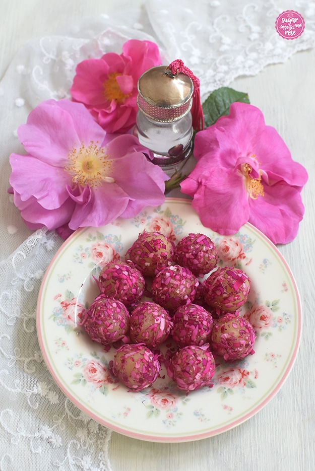 Ein kleiner Teller mit Rosendekor, darauf in Rosenblüten gerolltes Rosenmarzipan, dahinter drei unterschiedlich rosafarbene Blüten der Apothekerrose mit sichtbaren Staubgefäßen