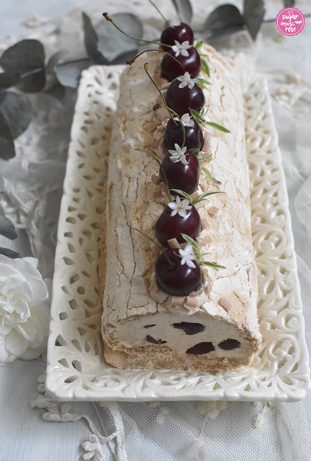 Kaffee-Baiserrolle auf Keramiketagere, dekoriert mit dunkelroten Kirschen  mit Stiel und kleinen weißen, sternförmigen Blüten und grünen Blättern